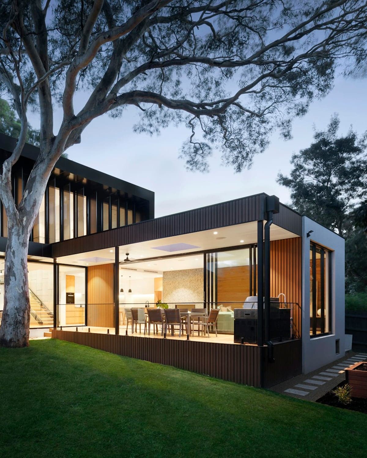 Casa moderna de madera y cristal iluminada al atardecer, con jardín y un árbol grande